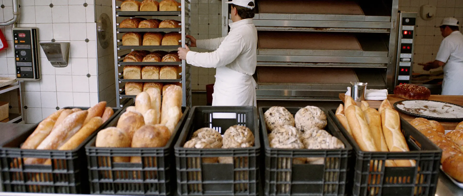 Padaria profissional em produção, com carrinho de bandejas de pães e caixas de pão fresco prontas para entrega